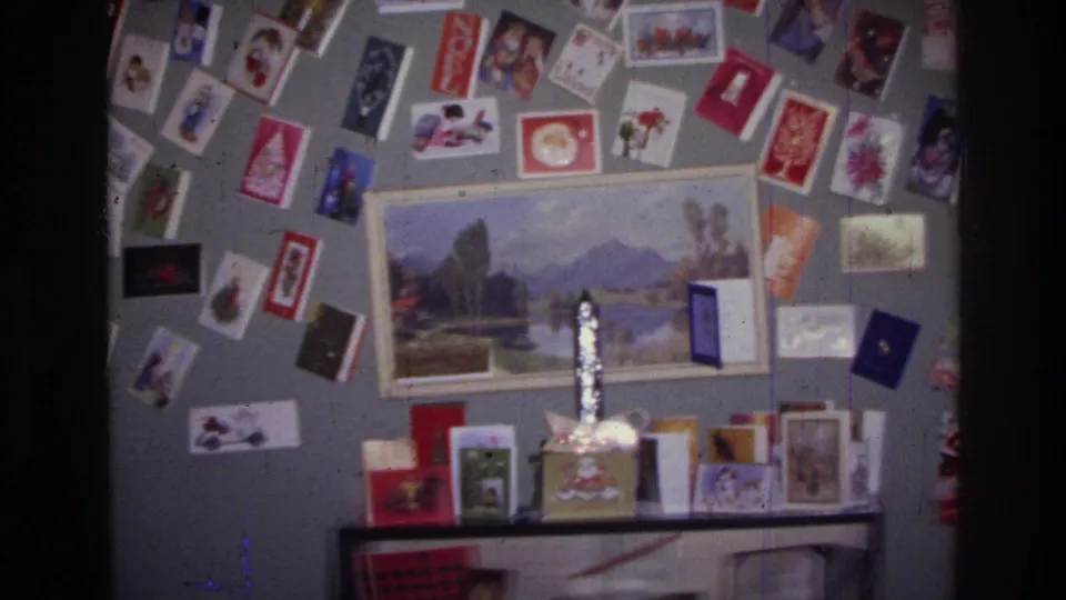 Harlem 1976 8mm frame showing colorful holiday cards around a framed lake-and-mountain print, with small display items on the shelf.