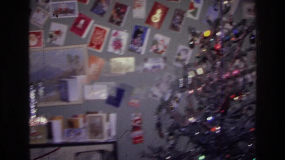1976 Harlem home-movie frame showing a silver Christmas tree near a gray wall filled with holiday greeting cards.