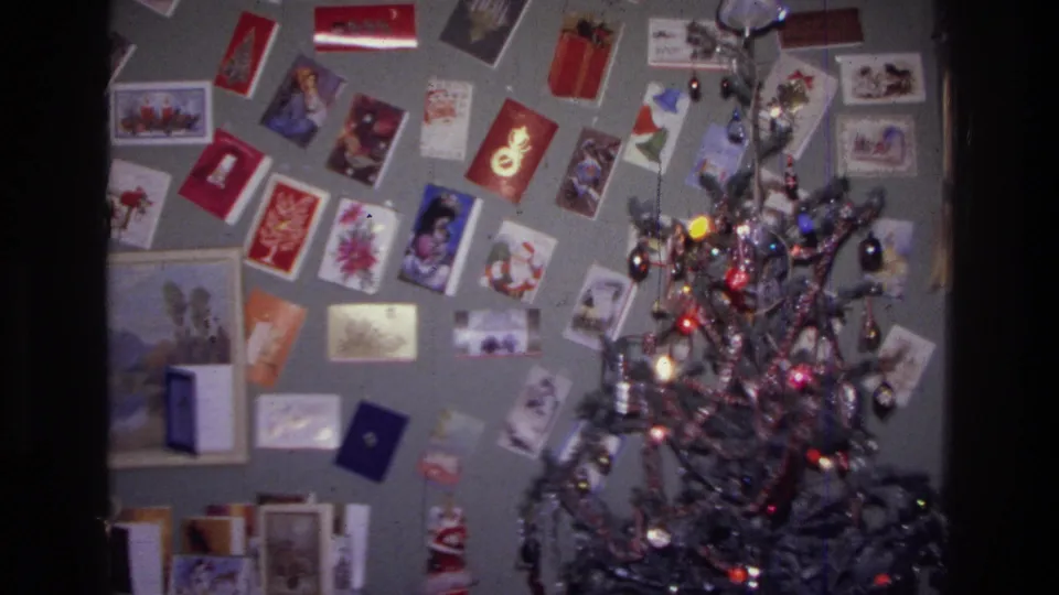 1976 Harlem home-movie view of a tinsel Christmas tree set against a gray wall covered with greeting cards.
