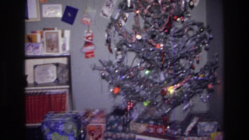 1976 Harlem home-movie frame showing a silver tinsel Christmas tree, ornaments, lights, and wrapped gifts in a private living room.