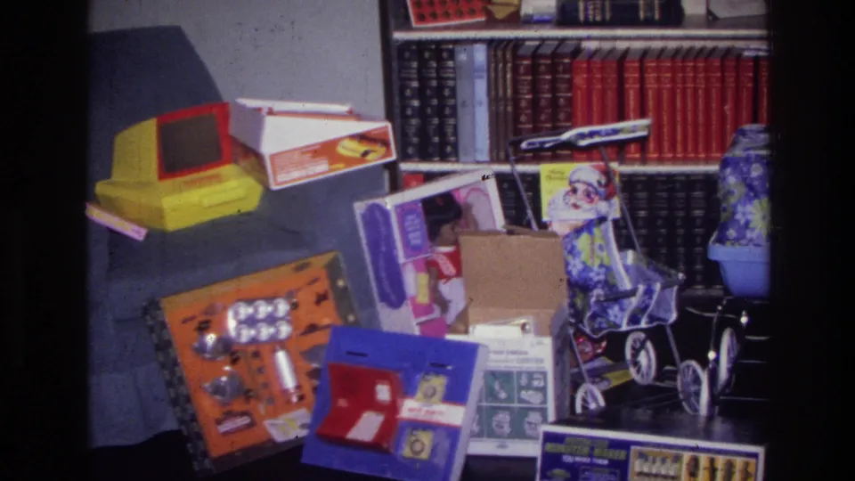 1976 Harlem home-movie frame with a toy stroller, boxed toys, gift packaging, and bookshelves in a private Christmas interior.