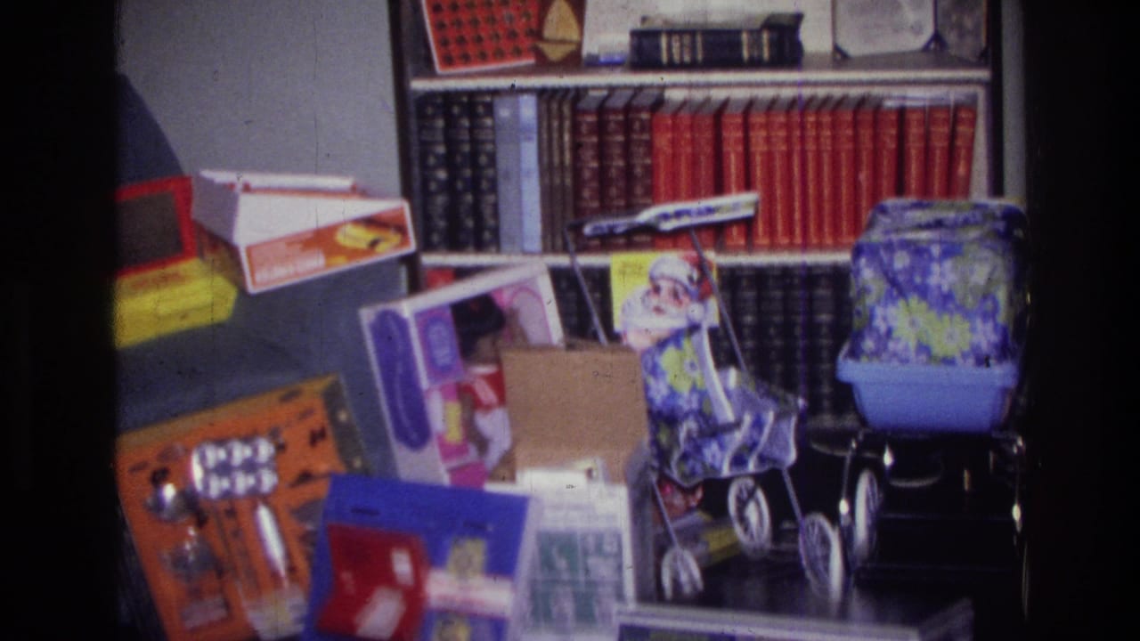 1976 Harlem home-movie frame showing toy boxes, a doll stroller, red-bound books, greeting cards, and holiday decorations in a private room.