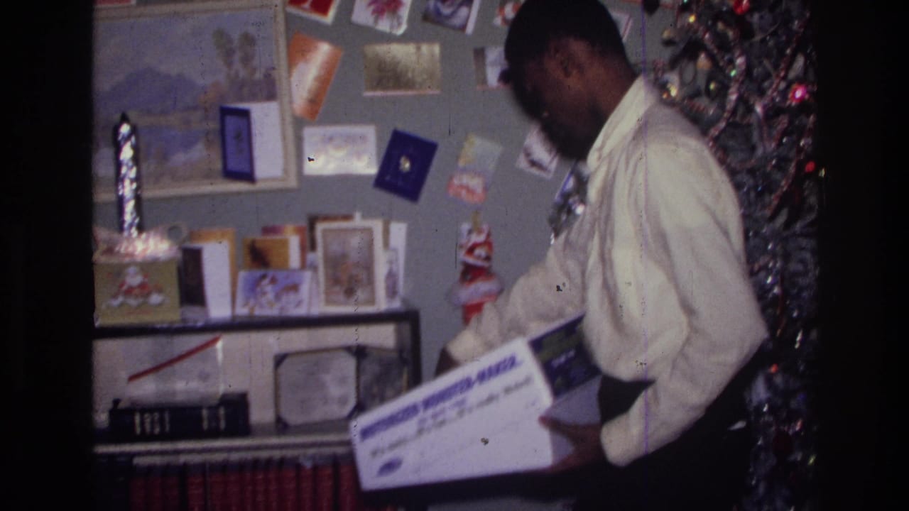 Person in a white shirt holds a boxed item beside shelves, cards, and a Christmas tree.