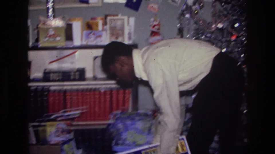 1976 Harlem home-movie frame showing a person bending toward the floor beside red-bound books, cards, and holiday decorations.