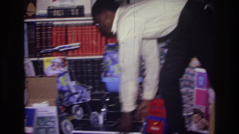 1976 Harlem home-movie frame showing a person lowering a boxed gift among toys, a doll stroller, bookshelves, and tinsel tree decorations.