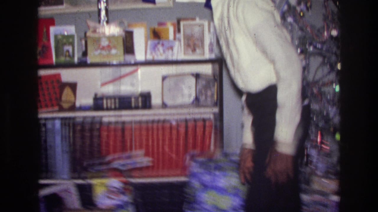 In a 1976 Harlem home interior, a man in a white shirt stands by shelves holding books, cards, and holiday objects.