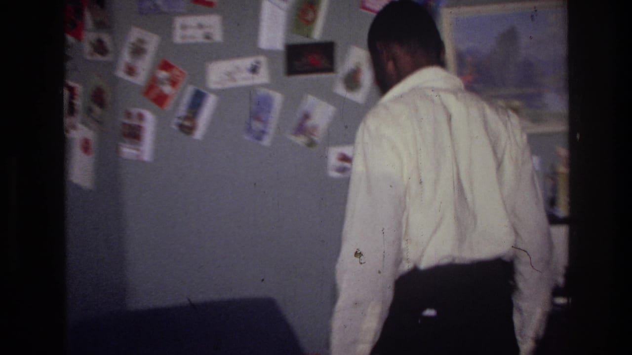 A man in a white shirt walks through a decorated Harlem room with cards and pictures pinned to the wall.