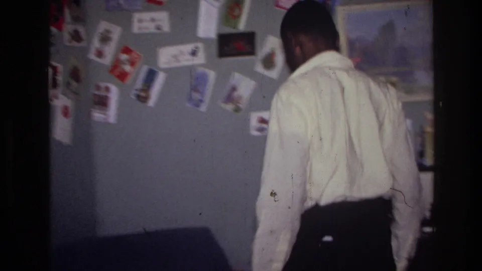 A man in a white shirt walks through a decorated Harlem room with cards and pictures pinned to the wall.