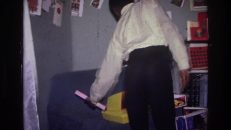 The man bends near a blue couch, holding small objects amid cards, shelves, and household decorations.