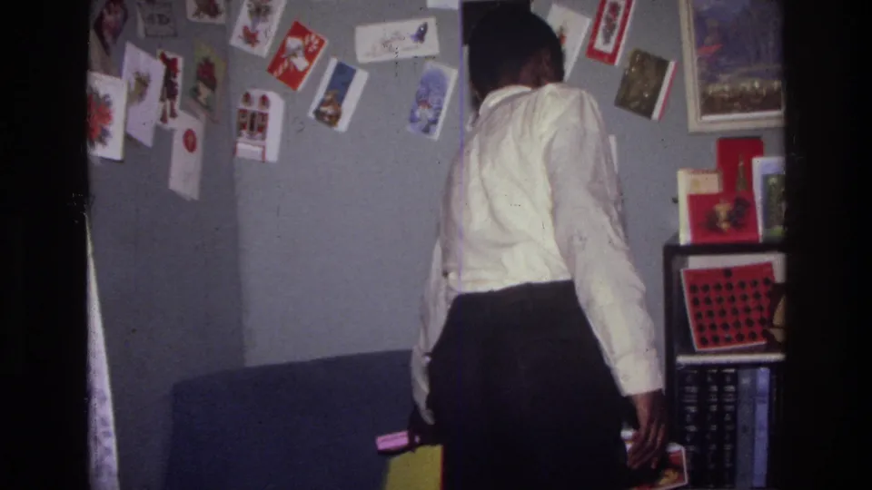 In the Harlem home interior, the man walks away with small items near a couch, card-covered wall, and shelves.