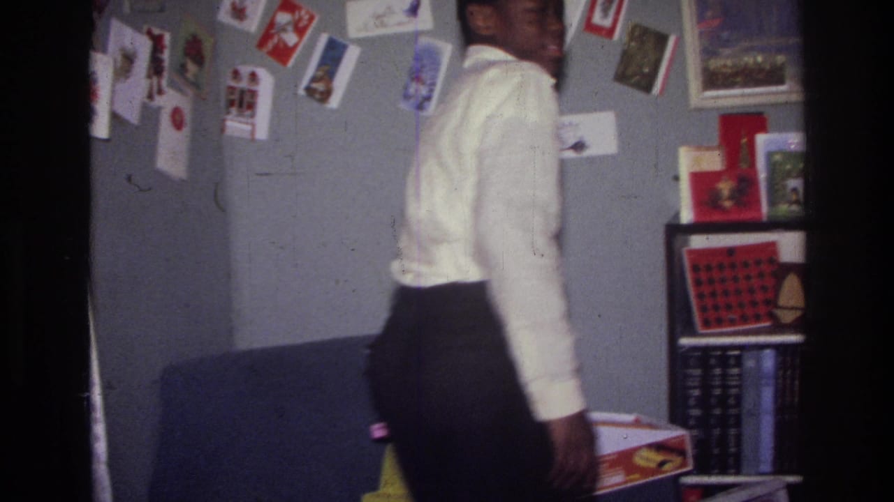 1976 Harlem home-movie frame showing a person in a white shirt near a wall covered with Christmas cards and framed pictures.
