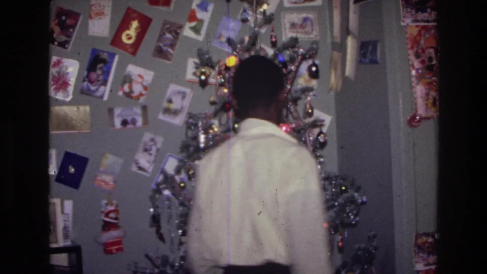 1976 Harlem home-movie frame showing the subject from behind near a lit Christmas tree and card-covered wall.