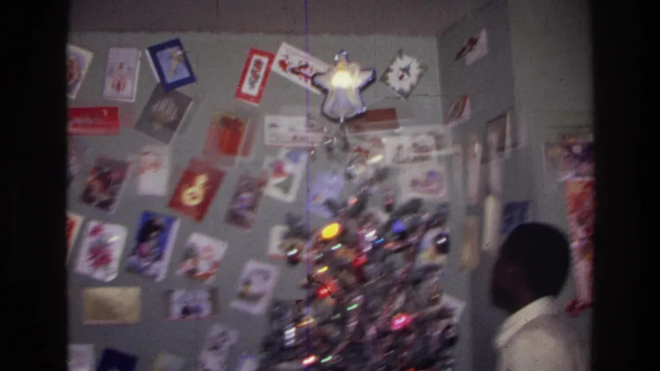 1976 Harlem interior frame with a glowing Christmas tree, wall cards, framed pictures, and the subject partly visible at right.