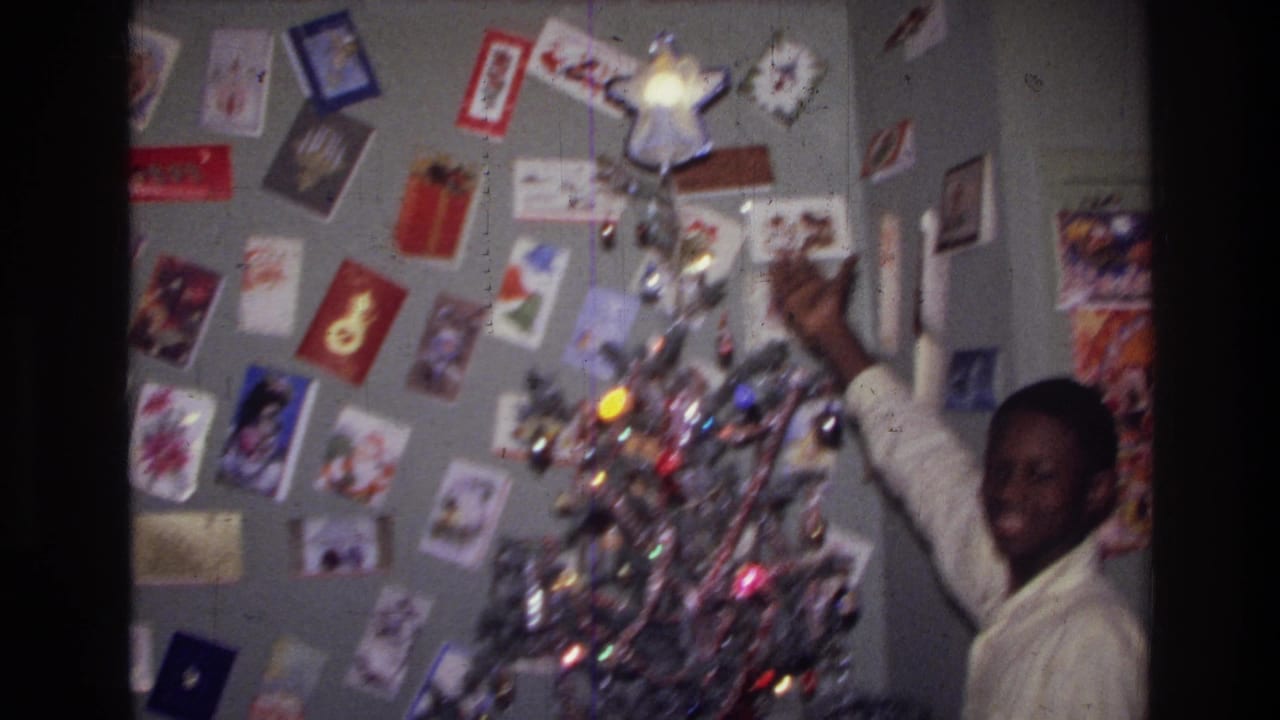 1976 Harlem home-movie frame of a teenager in a white shirt reaching to adjust a star atop a silver tinsel Christmas tree.