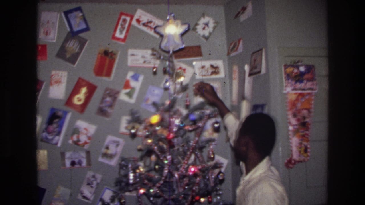 1976 Harlem home-movie frame showing a teenager beside a lit silver tinsel tree, with holiday cards on the wall behind him.