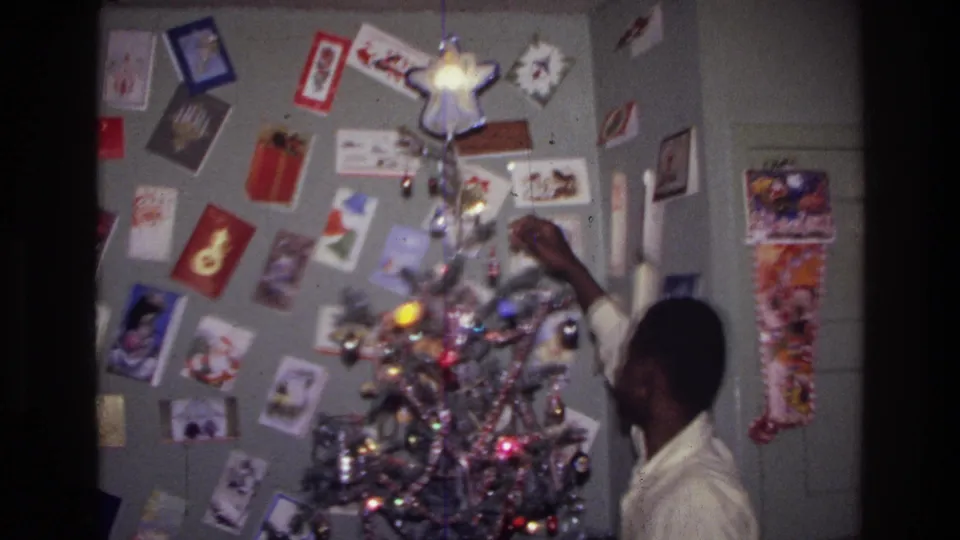 1976 Harlem home-movie frame showing a teenager beside a lit silver tinsel tree, with holiday cards on the wall behind him.