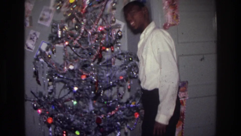 1976 Harlem home-movie frame of a smiling teenager standing next to a decorated silver tinsel Christmas tree.