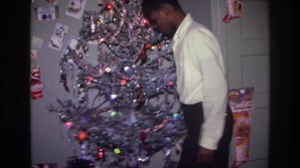 1976 Harlem home-movie frame showing a teenager looking down next to a lit silver tinsel Christmas tree in a decorated room.