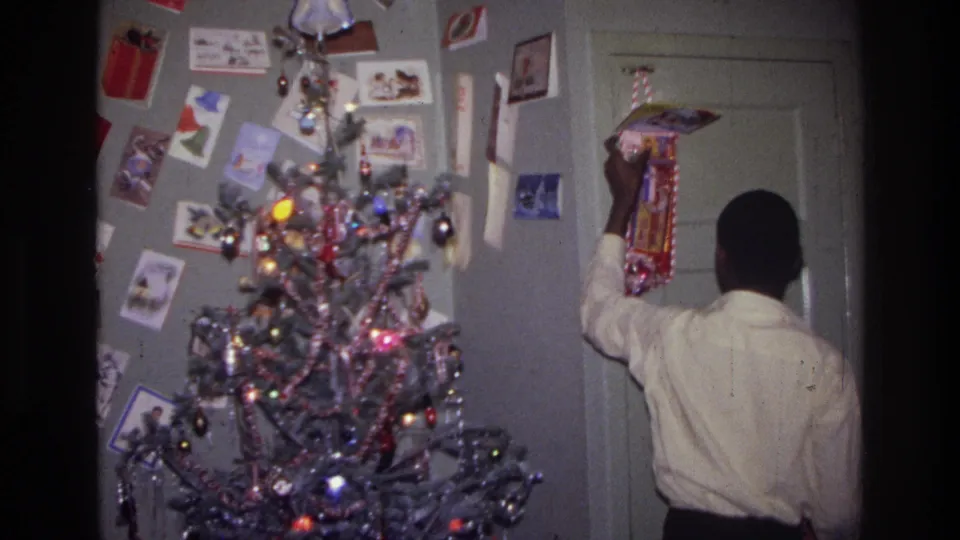 Home-movie frame from Harlem in 1976 with a person lifting a red decoration by the door near a lit Christmas tree.