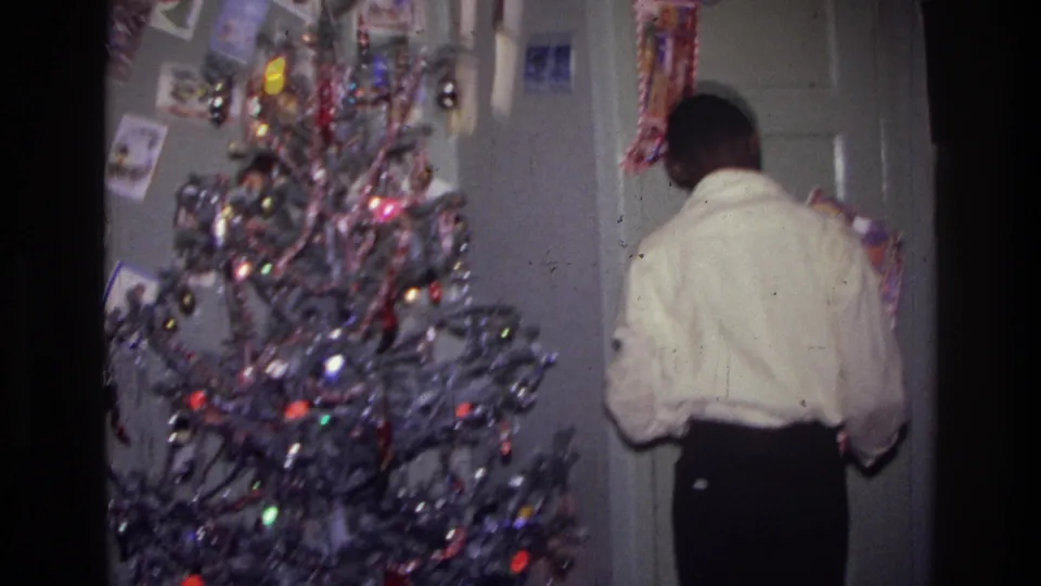 1976 Harlem holiday interior with a person turned toward the door and a decorated Christmas tree in the foreground.
