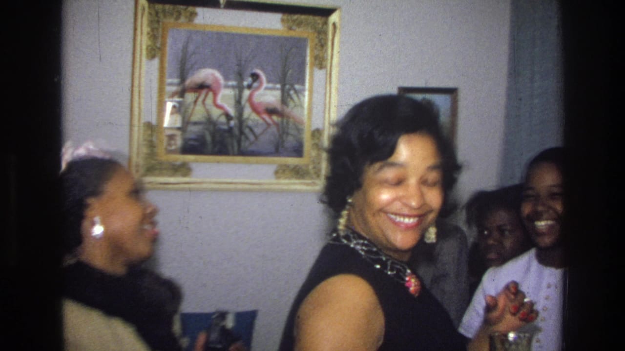 1976 Harlem home-movie frame of African American women laughing in a living room, with drinks, framed wall art, and soft 8mm grain.
