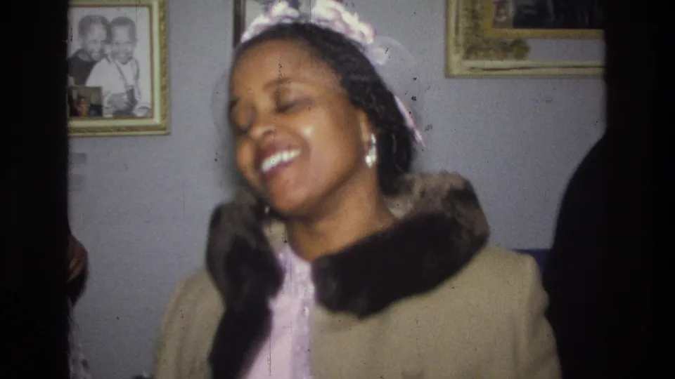 Close 1976 Harlem home-movie frame of a smiling woman in a beige coat with a dark collar, filmed inside a private living room.