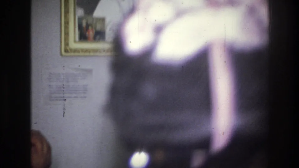 Soft, partially obscured 8mm frame from the 1976 Harlem interior gathering, with blurred foreground movement and framed wall decor behind it.