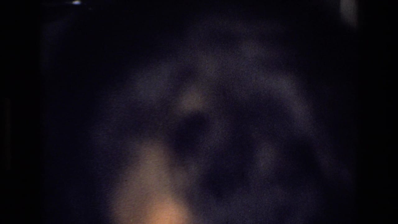 Partially blocked 8mm frame from the Harlem home-movie scene, with only a blurred foreground shape and dim interior light visible.