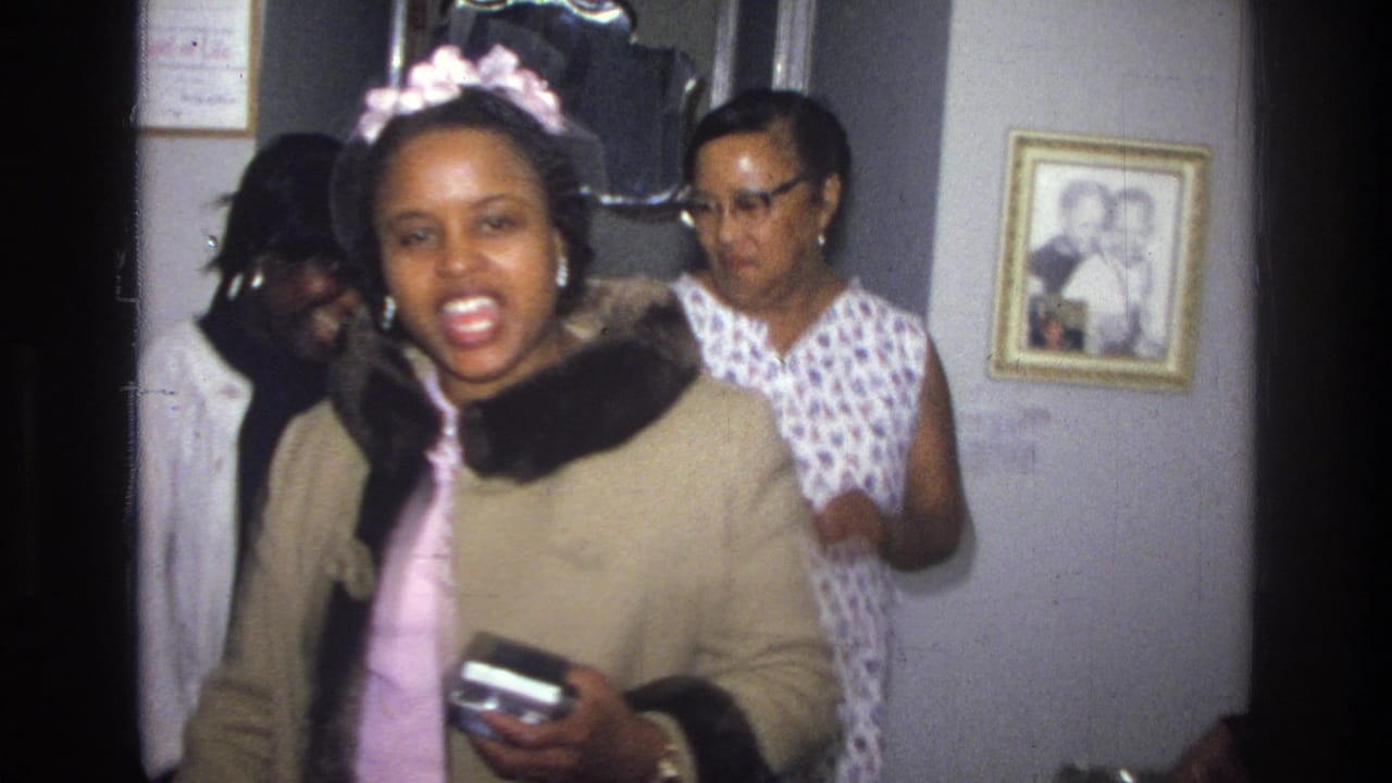 Young woman in a beige coat and pink headband stands indoors holding a small camera as others stand behind her.