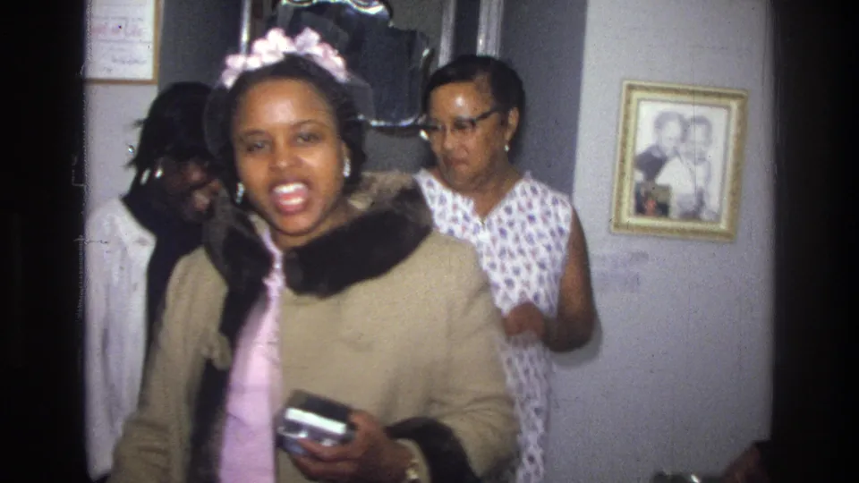 In the 1976 Harlem interior, a young Black woman in a beige coat and pink headband holds a small camera while family members stand nearby.