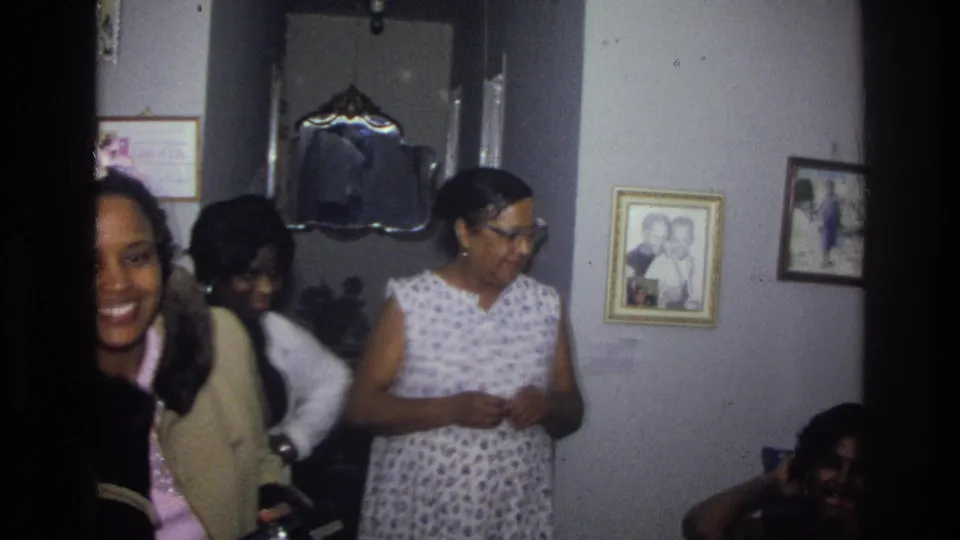 1976 Harlem home interior with several Black family members near framed photographs and a mirror as a woman smiles at the edge of the frame.