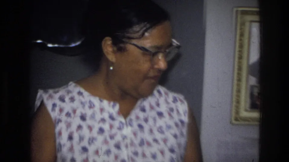 An older Black woman in glasses and a sleeveless floral dress stands in profile in the 1976 Harlem home interior, with framed pictures nearby.