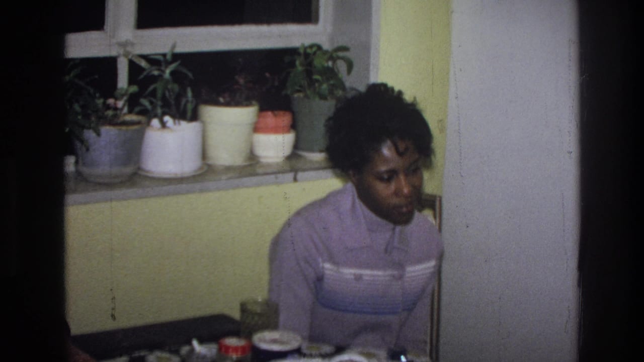 In 1976 Harlem home-movie footage, a woman in a striped purple sweater sits at a table near a yellow wall and potted plants.