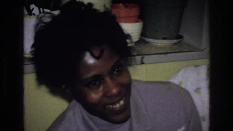The woman turns toward the handheld 8mm camera and smiles, with shelves and a yellow wall behind her.