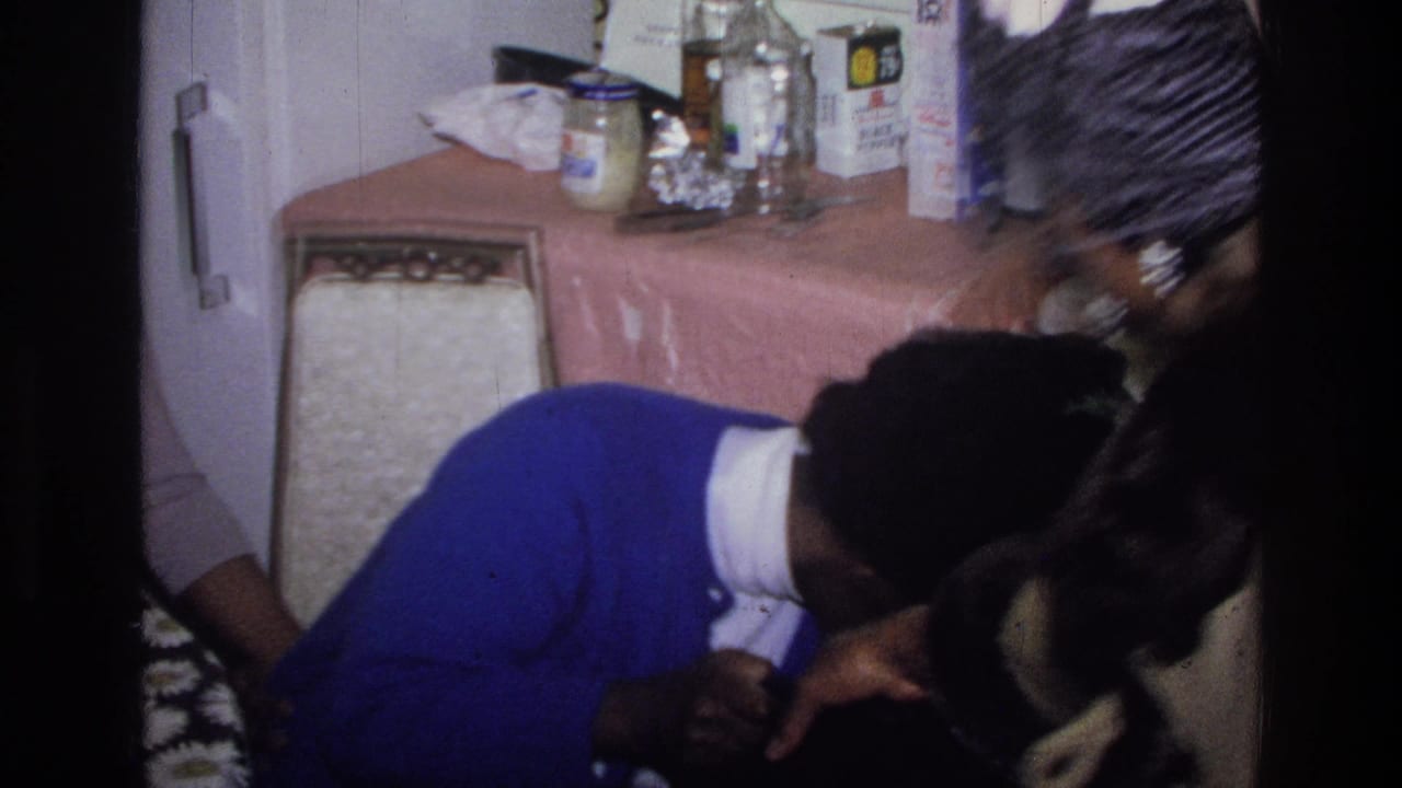 In a 1976 Harlem home-movie frame, a woman in a blue sweater leans forward beside another adult in a compact kitchen.