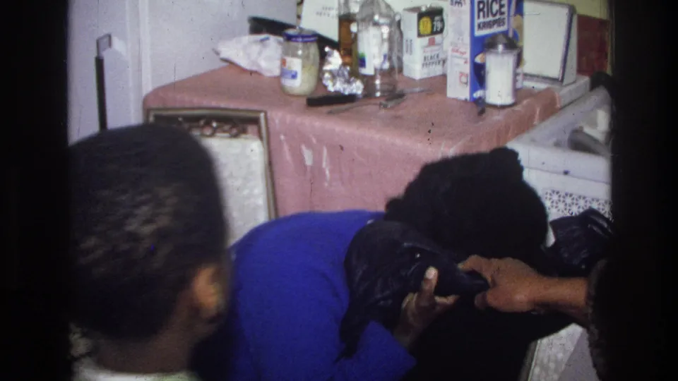Two adults appear close together in a modest Harlem kitchen, with a refrigerator, jars, and boxed pantry items visible behind them.