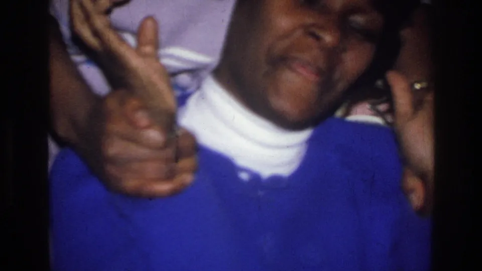 A woman in a blue sweater and white turtleneck smiles and gestures toward the camera in a 1976 Harlem kitchen home movie.
