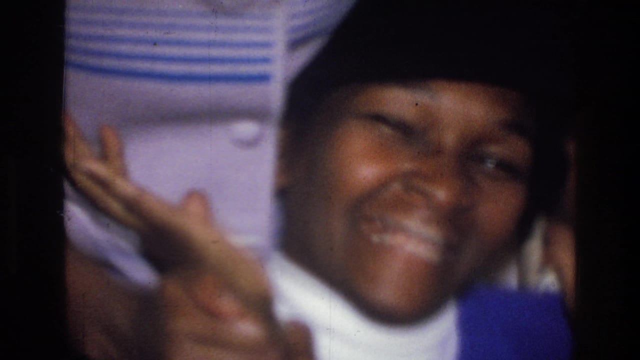 In 1976 Harlem home-movie footage, a woman in a blue sweater and black knit hat smiles and gestures close to the camera indoors.