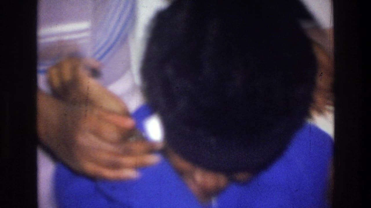 In the Harlem interior, the woman in blue lowers her head as nearby hands and other people crowd the close handheld frame.