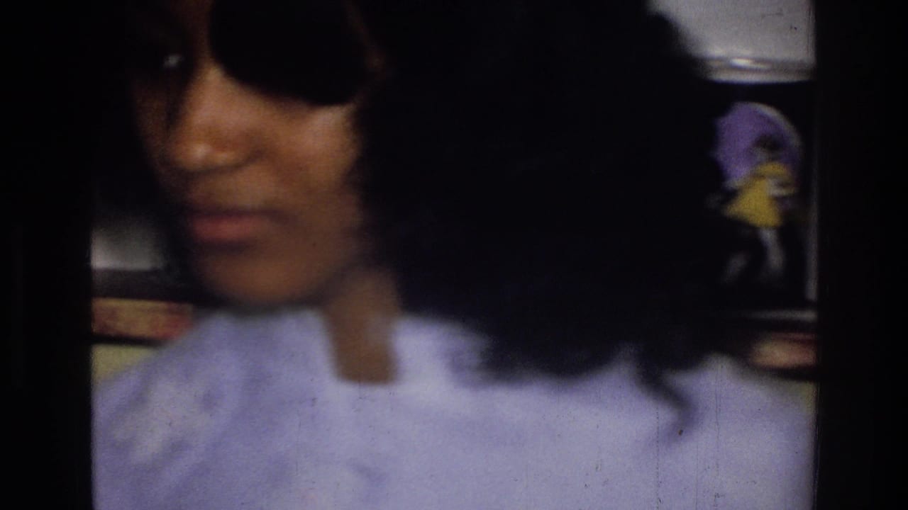 In the supplied 1976 Harlem home-movie context, a woman with dark hair and sunglasses appears in soft focus inside a room.