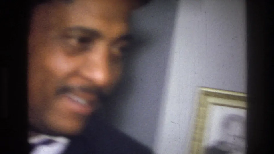 A smiling man with a mustache faces the camera in the supplied Harlem home-movie setting, with a framed wall picture nearby.