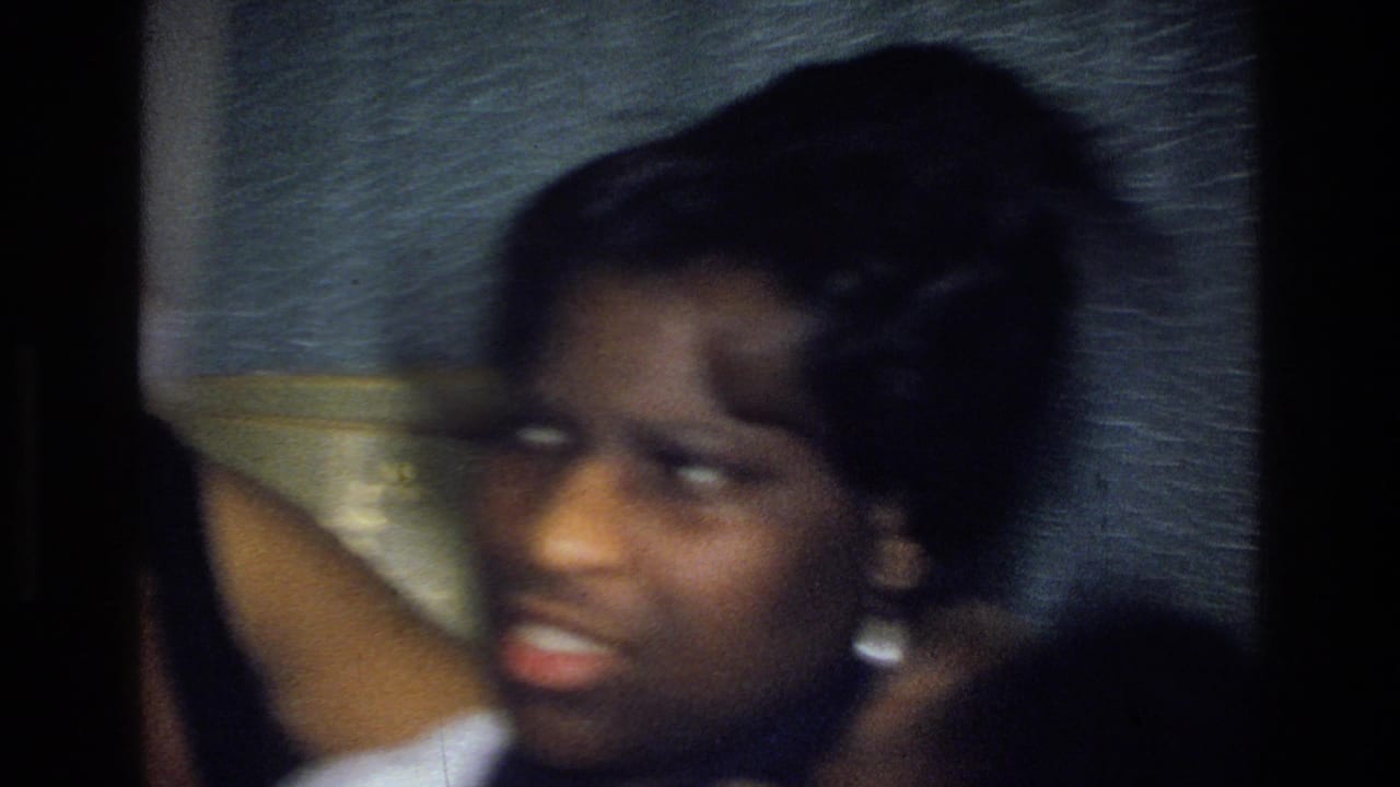 Soft-focus 1976 Harlem home-movie frame of a young person indoors, wearing silver earrings as an arm crosses the foreground.