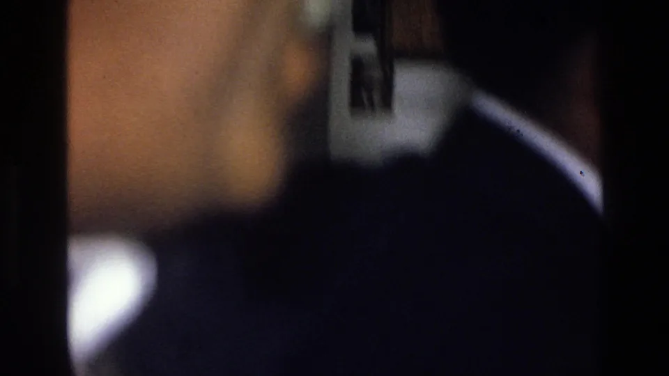 Soft 8mm frame from the 1976 Harlem clip with a close partial face and dark-clothed figure in a low-light interior.