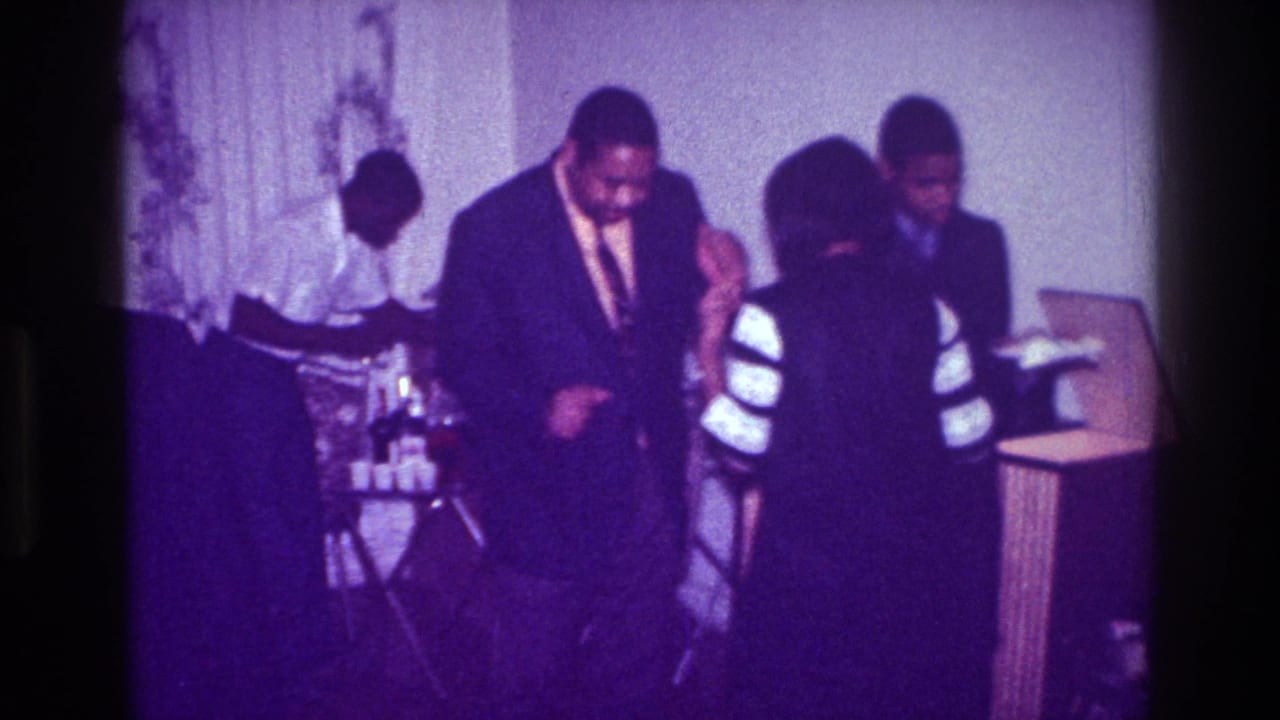 Adults in formal clothing dance in a softly lit room with curtains and a table nearby.