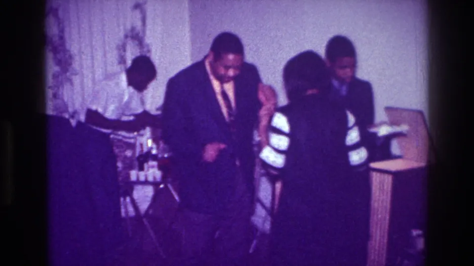 1976 Harlem home-movie frame showing a suited man and a woman with white striped sleeves dancing at an indoor celebration.