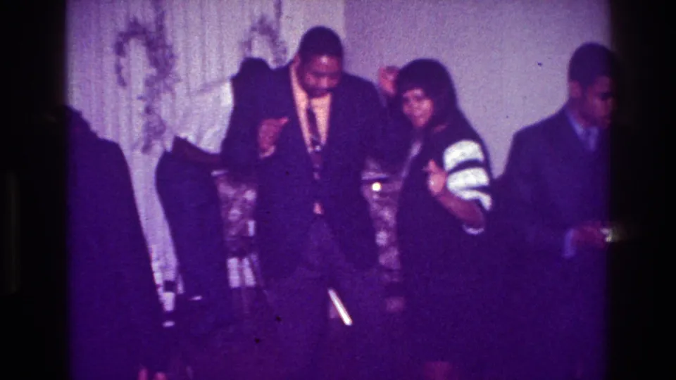 1976 Harlem party scene with adults dancing in formal attire inside a private room with curtains and soft purple film color.
