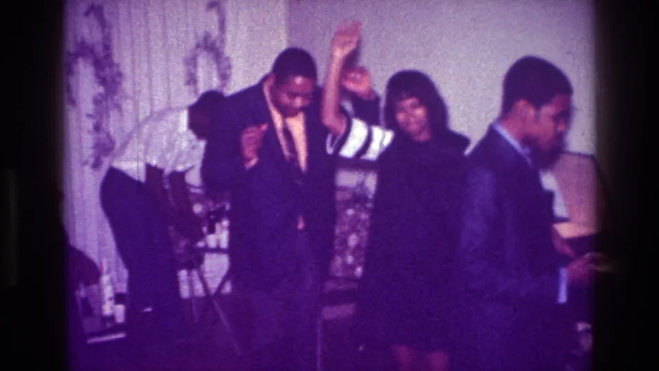 1976 Harlem indoor celebration with a woman raising her arm as she dances near a suited man and guests in formal clothing.