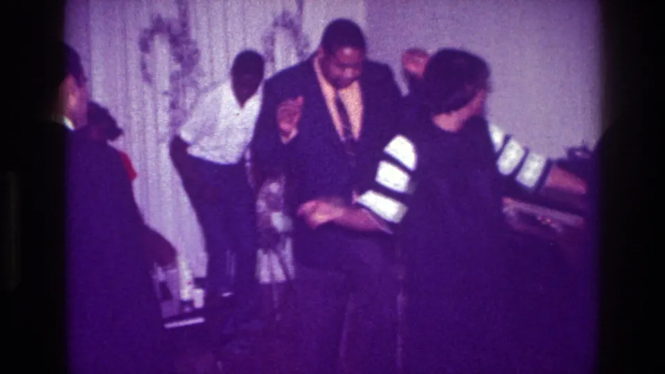 1976 Harlem home-movie frame showing a woman in striped sleeves turning during a dance, with suited guests and a curtained interior behind her.