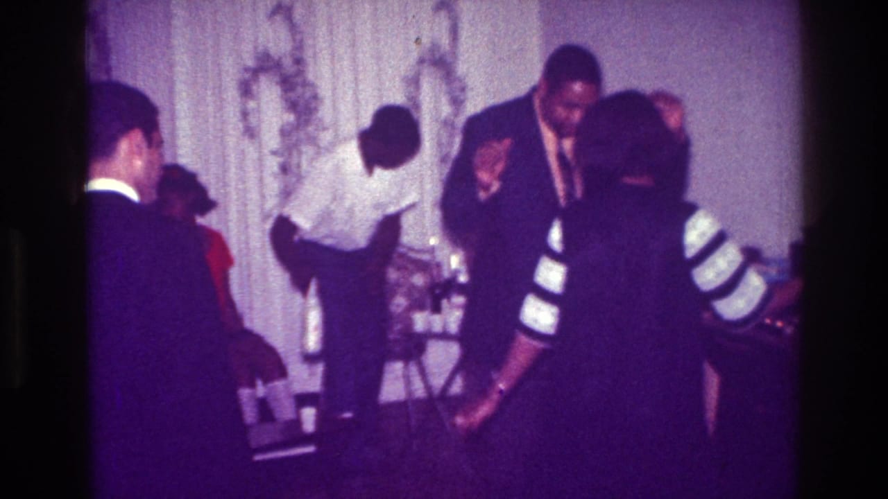 1976 Harlem home-movie frame showing suited guests, a woman in a striped dress, and a man near a bottle at an indoor party.