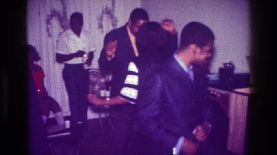 1976 Harlem home-movie frame with a smiling suited man crossing in front of dancers and a drinks table in a private interior.